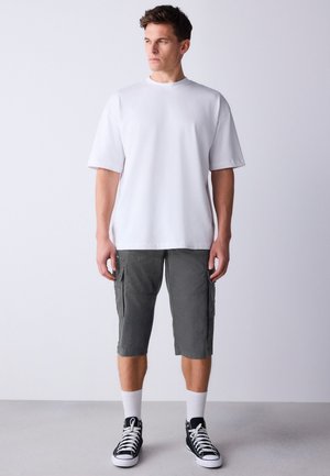 Giovane uomo che indossa una maglietta bianca semplice, pantaloncini cargo grigi, calzini bianchi e sneakers alte nere, in piedi su sfondo bianco.