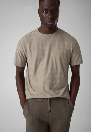 Homme portant un t-shirt beige à manches courtes avec un motif floral subtil et un pantalon vert olive, debout les mains dans les poches sur fond gris.