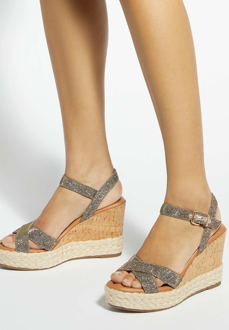 Piernas con sandalias de cuña plateadas brillantes con tiras, tacones de corcho y detalles de cuerda trenzada en las suelas de la plataforma.