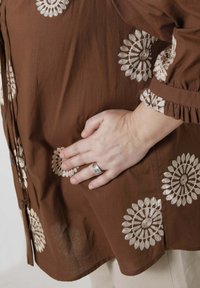 Bruine katoenen blouse met witte geborduurde bloemmotieven, knoopsluiting en ruches aan de mouwen. Hand rustend op de stof.