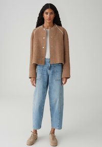 Donna con capelli lunghi e ricci indossa una giacca marrone texturizzata, una maglia bianca, jeans a vita alta di colore azzurro chiaro e sneakers beige, in piedi su uno sfondo grigio.