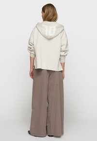 Sudadera gris claro con un gran bolsillo frontal y capucha, que presenta el número "10" en blanco en la parte posterior. Combinada con pantalones beige de corte holgado.