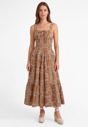 Kobieta stojąca ubrana w brązową, wzorzystą sukienkę midi bez rękawów ze ściągaczem w talii i beżowe koturny espadryle z wiązaniami wokół kostek.