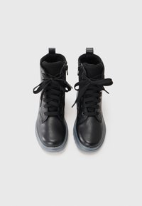 Par de botas de cuero negro hasta el tobillo con cordones anudados en forma de lazo, cremallera lateral y suelas de goma gris sobre fondo blanco.