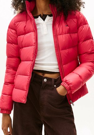 Mujer con una chaqueta acolchada roja brillante sobre una camiseta blanca corta, combinada con pantalones de pana marrón oscuro de talle alto.