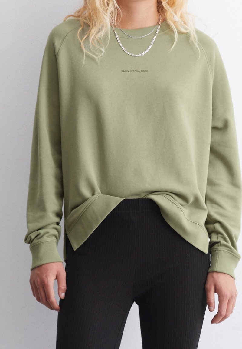 Olivově zelená mikina s kulatým výstřihem, žebrovanými manžetami a lemem. Obsahuje jemné brandování na hrudi. Spárovaná s černými žebrovanými kalhotami.