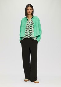 Gequilte mintgroene jas met knopen, blouse met geometrisch patroon, zwarte wijde broek en witte en tan sneakers.