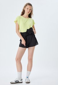 Hellgrünes Kurzarmoberteil, schwarze Shorts mit gebundenem Bund, weiße Sneakers mit schwarzen Streifen und weiße strukturierte Socken.