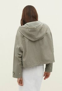 Vrouw met lang bruin haar in een lichtgrijs cropped jack met capuchon en witte broek, staande met haar rug naar de camera.