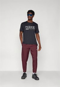 Nike svart t-skjorte med hvit logo, burgunder avsmalnede bukser og svarte joggesko. Modellen har også på seg blå solbriller. Enkel atletisk design.