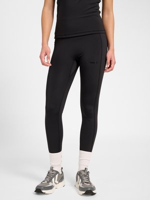 Osoba ubrana w czarne legginsy sportowe, białe skarpetki i szaro-białe buty sportowe, stojąca na jednolitym tle.