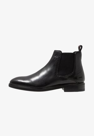 Bottines Chelsea en cuir noir avec une surface lisse, des panneaux latéraux élastiques, des perforations décoratives et une languette pour un enfilage facile.