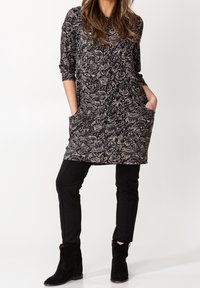 Nainen, joka on pukeutunut mustavalkoiseen paisley-kuvioiseen tunikaan, jossa on taskut, mustiin slim-fit-housuihin ja mustiin nilkkureihin, seisoo valkoista taustaa vasten.