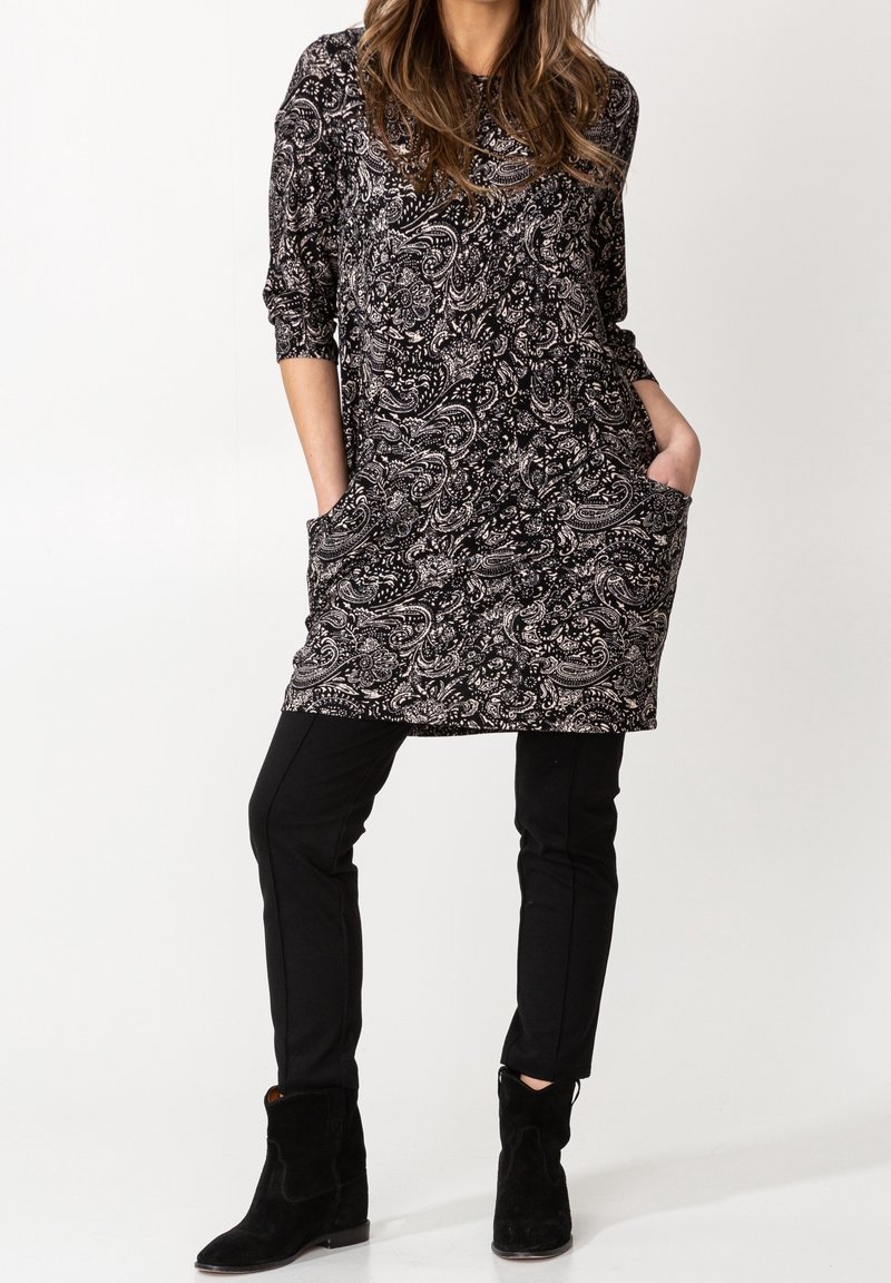 Nainen, joka on pukeutunut mustavalkoiseen paisley-kuvioiseen tunikaan, jossa on taskut, mustiin slim-fit-housuihin ja mustiin nilkkureihin, seisoo valkoista taustaa vasten.