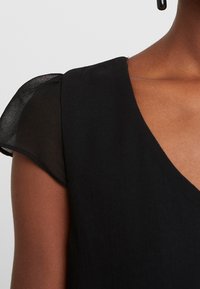 Svart klänning med korta, genomskinliga ärmar som fladdrar, en V-ringning och en slät texturerad byst. Tyget verkar lätt och flödande.