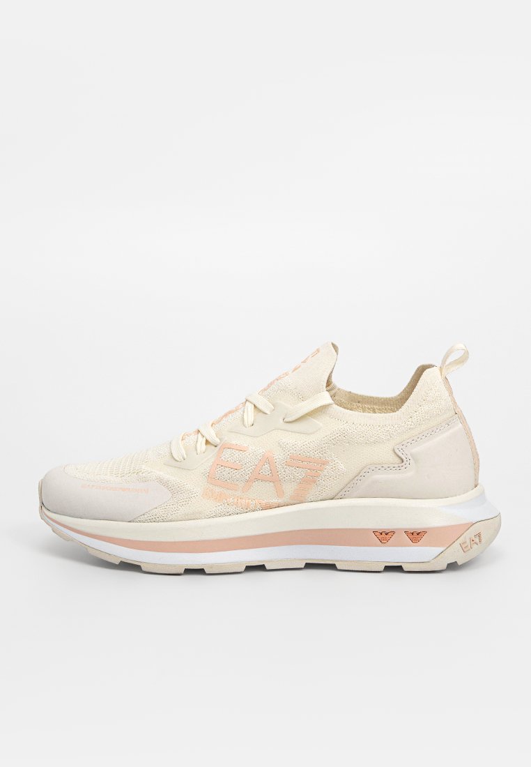 Zapatilla deportiva beige con parte superior de punto texturizado, acentos rosas, suela gruesa y detalles de logo visibles en el lateral y en la parte trasera.