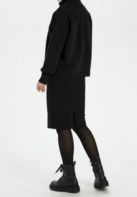 Mujer vestida con blusa negra de manga larga, falda hasta la rodilla, medias negras y botas negras con cordones, de pie contra un fondo liso.