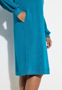 Kobieta w niebieskiej sukience z długim rękawem, z jedną ręką w bocznej kieszeni, stojąca na białym tle.