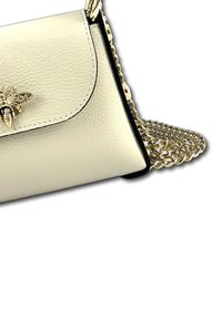 Lichtbeige leren handtas met een klepafsluiting, gouden bij-embleem en een gestructureerde kettingriem. Heeft een zwarte rand langs de randen.