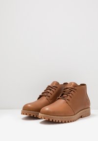 Botas de cuero marrón hasta el tobillo con punteras redondeadas, cordones redondeados y suelas de goma texturizada. Presentan un detalle de logo lateral y un acabado suave.