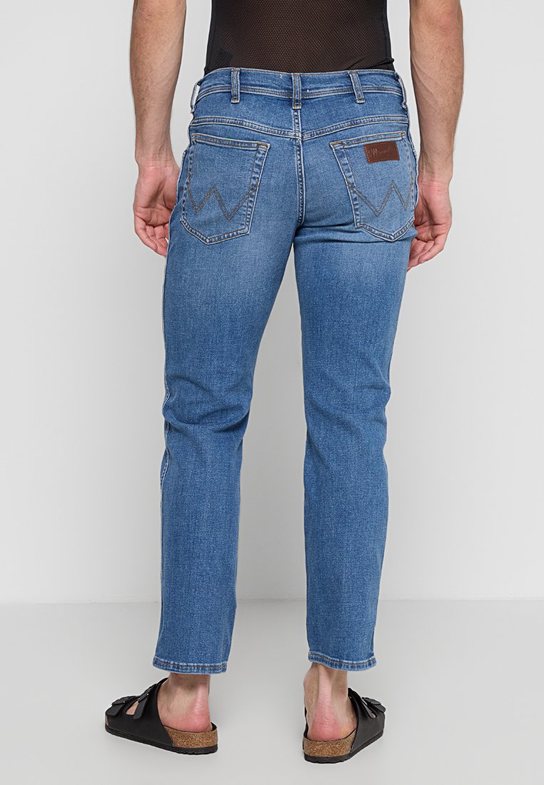 Osoba stoi tyłem w niebieskich dżinsach, ukazując tylne kieszenie i brązową skórzaną łatkę, w połączeniu z czarnymi sandałami. Białe tło.