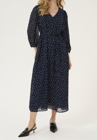 Robe longue à manches longues bleu marine avec des pois blancs, décolleté en V, taille cintrée et jupe fluide. Le modèle porte des mules à talons noires.