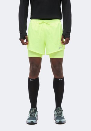 Homme portant un short de course superposé jaune néon, un haut noir à manches longues, des chaussettes noires Nike montant jusqu'aux genoux et des chaussures de course grises.