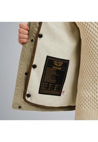 Khaki bunda s béžovým štruktúrovaným podšívkou. Obsahuje čiernu štítku na bielom filci so špecifikáciami, zabezpečenú tmavými gombíkmi.