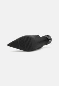 Chaussure noire à talon avec un bout pointu, dessus lisse et semelle en caoutchouc arborant des rainures horizontales pour une meilleure adhérence.