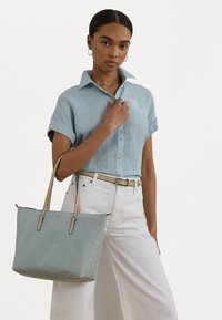 Bolsa tote azul clara com alças bege, textura suave, forma retangular e logotipo prateado. Modelo veste uma camisa azul clara e calças brancas.