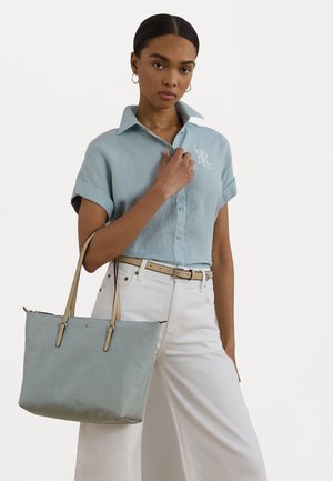 Lichtblauwe schoudertas met beige handvatten, gladde textuur, rechthoekige vorm en zilveren logo. Model draagt een lichtblauw shirt en witte broek.