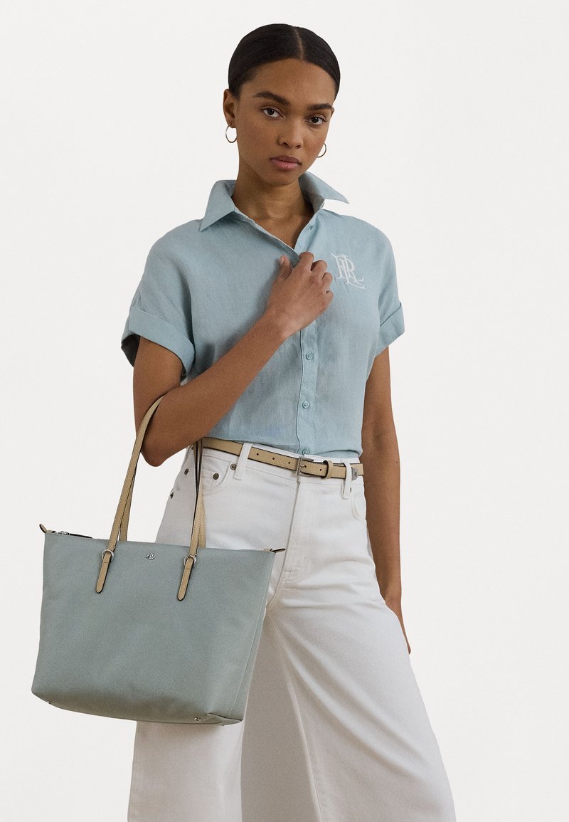 Bolsa tote azul clara com alças bege, textura suave, forma retangular e logotipo prateado. Modelo veste uma camisa azul clara e calças brancas.