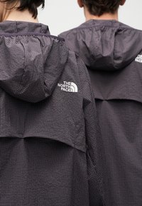 Δύο άτομα που φορούν μαύρα καρό μπουφάν The North Face με κουκούλες, εμφανίζονται από πίσω μπροστά σε απλό φόντο.