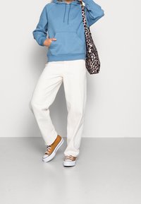 Persona che indossa una felpa blu, pantaloni bianchi, sneakers color senape, con una borsa a tracolla leopardata, in piedi con una mano in tasca.