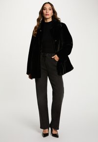 Cappotto in faux fur nero con colletto, doppi bottoni e maniche larghe, abbinato a jeans neri a vita alta e décolleté nere.