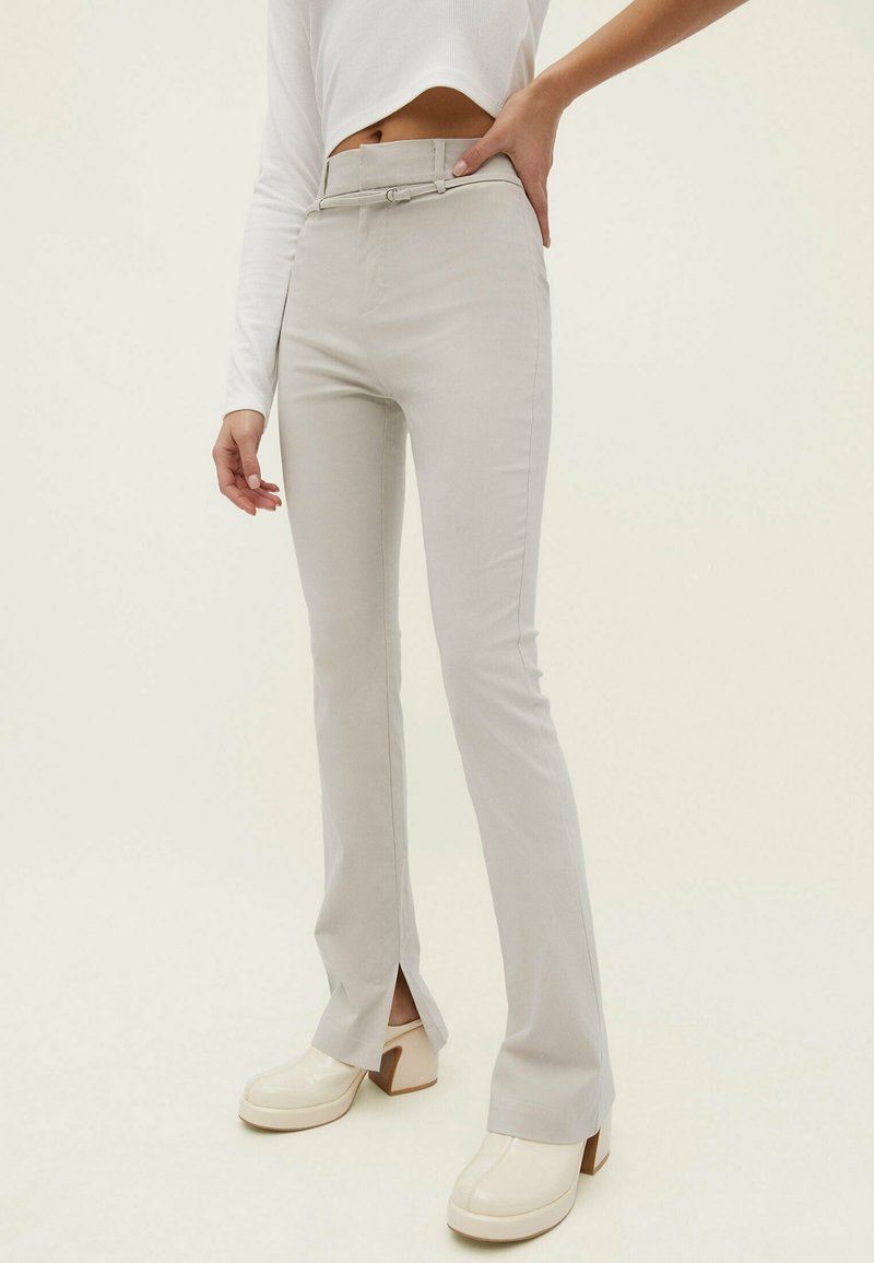 Femme portant un pantalon ajusté gris clair avec des fentes à l'avant, associé à un crop top blanc à manches longues et des chaussures à plateforme blanches.