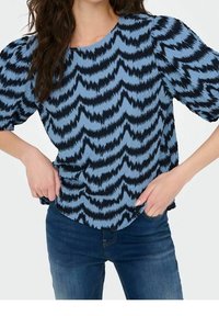 Blå topp med mörkt zigzag-mönster, korta puffärmar, rund halsringning och lös passform, i kombination med mörkblå jeans. Strukturerad tygdetalj.