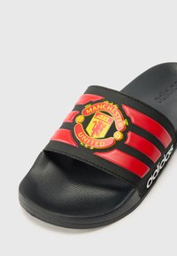 Black slide sandal with red-striped strap featuring a Manchester United logo, textured footbed, and smooth outsole.