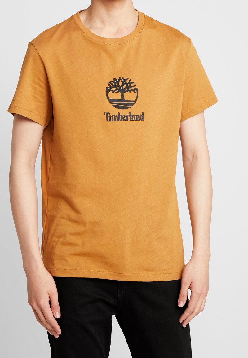 T-shirt en coton jaune moutarde avec un col rond. Présente un logo Timberland noir et un motif d'arbre à l'avant. Manches courtes, coupe décontractée.
