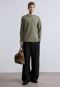 Πράσινο πουκάμισο με μακριά μανίκια και μικρό λογότυπο, συνδυασμένο με φαρδιά μαύρη παντελόνια και καφέ υφασμάτινη τσάντα με φερμουάρ και λουριά.