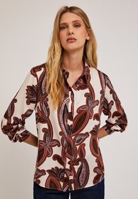 Bluză cu butoni de culoare crem, cu un model floral mare, îndrăzneț, în nuanțe de burgund și negru. Mâneci lungi, suprasolicitate. Material neted, cu guler.