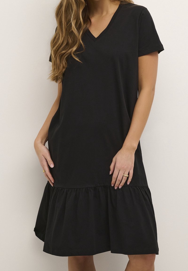 Femme portant une robe noire ample à manches courtes, col en V et ourlet à volants, debout devant un fond uni clair.