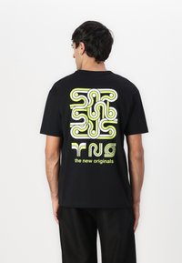 Svart bomulls t-skjorte med et dristig, abstrakt design i grønt og hvitt på baksiden, med teksten "de nye originalene" nederst.