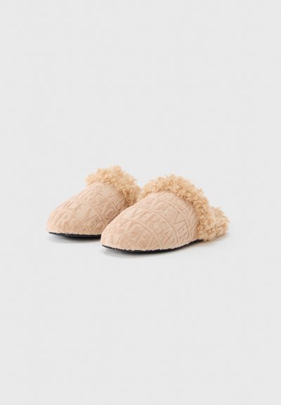 Paire de chaussons beiges à enfiler avec des lettres texturées et une bordure duveteuse autour de l'ouverture sur un fond blanc uni.