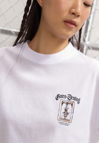 Tricou alb cu guler rotund, având un imprimeu grafic al unei cărți de tarot etichetate "Porțile Destinului" și "Omul Spânzurat" în negru și auriu.