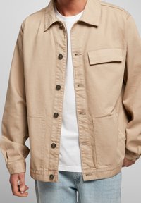 Beige canvasjacka med knappstängning framtill, spetsig krage och bröstficka. Har långa ärmar och en avslappnad passform. Bärs över en vit skjorta.