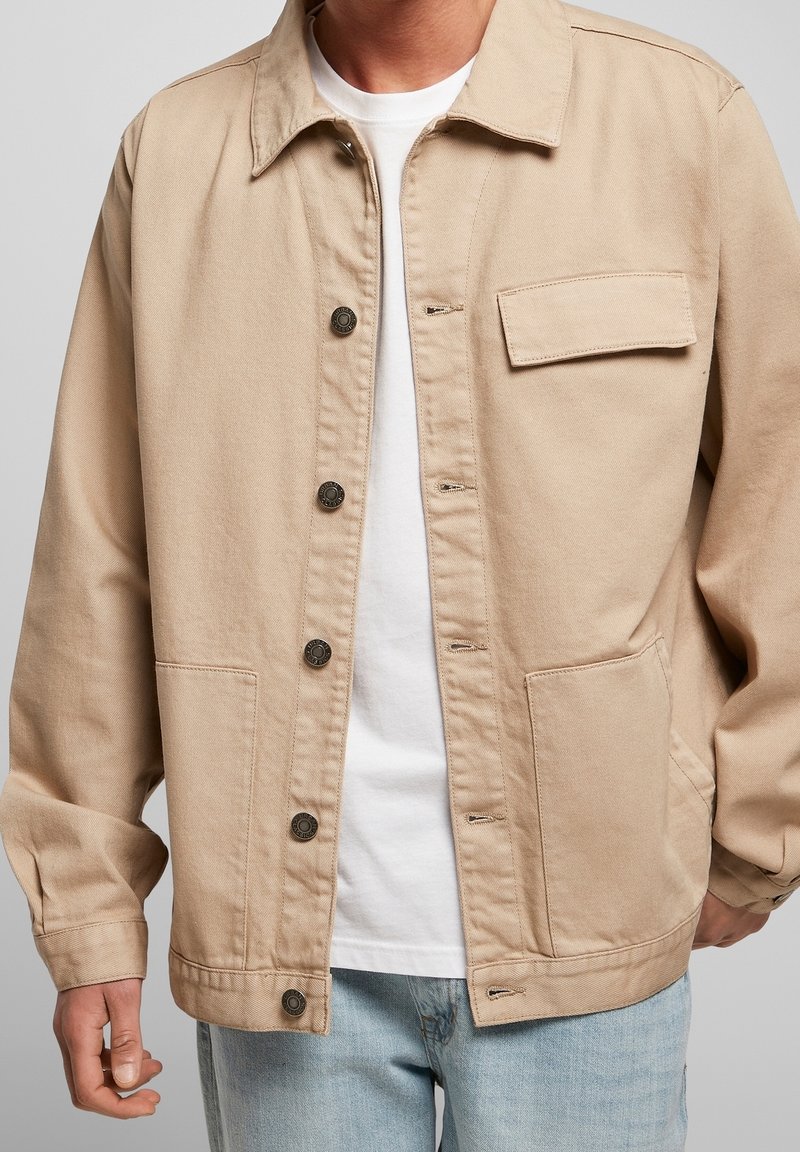 Beige canvasjacka med knappstängning framtill, spetsig krage och bröstficka. Har långa ärmar och en avslappnad passform. Bärs över en vit skjorta.