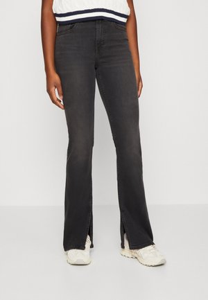 Person trägt schwarze Jeans mit geradem Bein und vorderen Knöchelschlitzen, weiße Turnschuhe und ein weißes ärmelloses Stricktop.