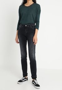 Dunkelgrünes Langarmoberteil mit einem mit Spitze verzierten Ausschnitt, kombiniert mit schwarzen Skinny-Jeans und schwarz-weißen Sneakers. Ein einfacher und lässiger Look.