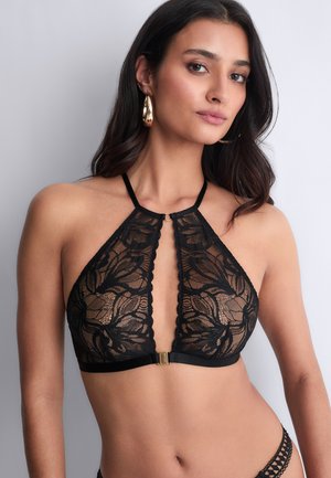 Femme portant un soutien-gorge halter en dentelle noire avec un motif floral et un fermoir en or, associé à un bas en dentelle noire assorti, posant contre un fond blanc.
